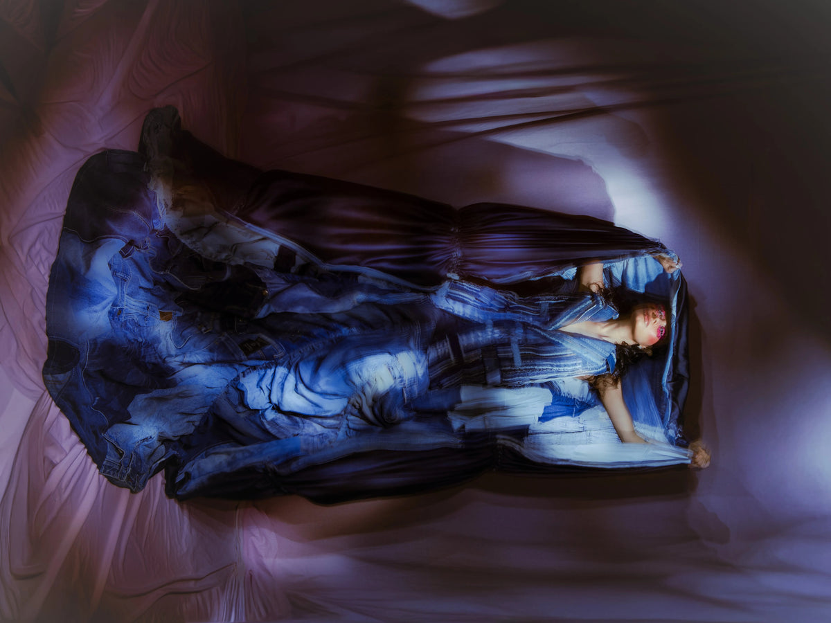 A woman stands in a dramatic studio setting wearing a voluminous blue and white draped gown or fabric ensemble, holding the material up above her head. The scene is lit with purple and blue theatrical lighting against a dark draped backdrop.