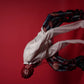 A person in a white shirt and ripped black-and-red pants crouches on a solid red studio background, with a red mesh head covering obscuring the face.