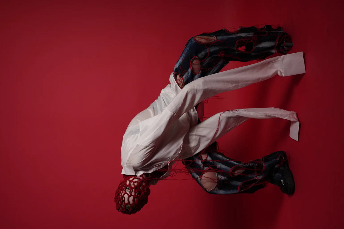 A person in a white shirt and ripped black-and-red pants crouches on a solid red studio background, with a red mesh head covering obscuring the face.