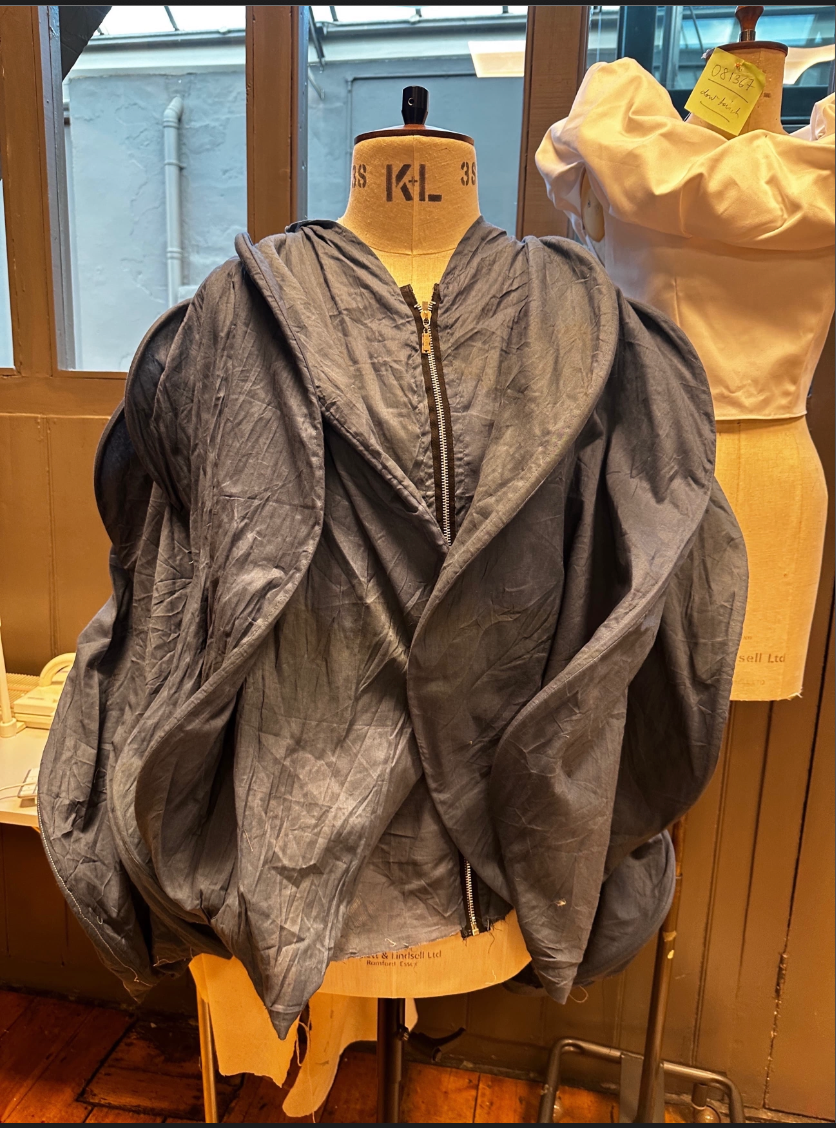A crinkled dark gray lightweight jacket displayed on a dress form mannequin, with a second mannequin wearing a cream puff-sleeve top in the background near large windows.