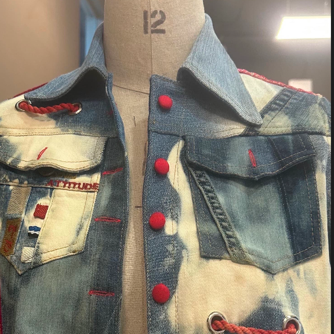 A cropped denim jacket displayed on a dress form mannequin. The jacket features a mix of light and dark blue denim panels with a bleached off-white center panel, red buttons, red drawstring details, and distressed frayed edges.